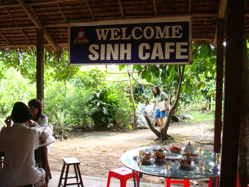 Một điểm nghỉ chân mái lá với biển hiệu 'Welcome Sinh Cafe' và bàn tròn bày thức ăn ở Đồng bằng sông Cửu Long.