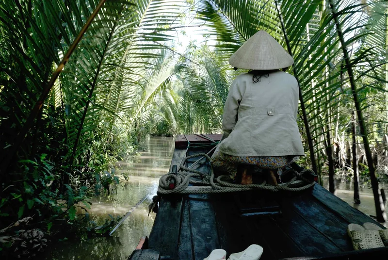 Người chèo đội nón lá ngồi ở mũi thuyền gỗ, lướt qua con rạch hẹp rợp bóng cây dừa ở Đồng bằng sông Cửu Long.