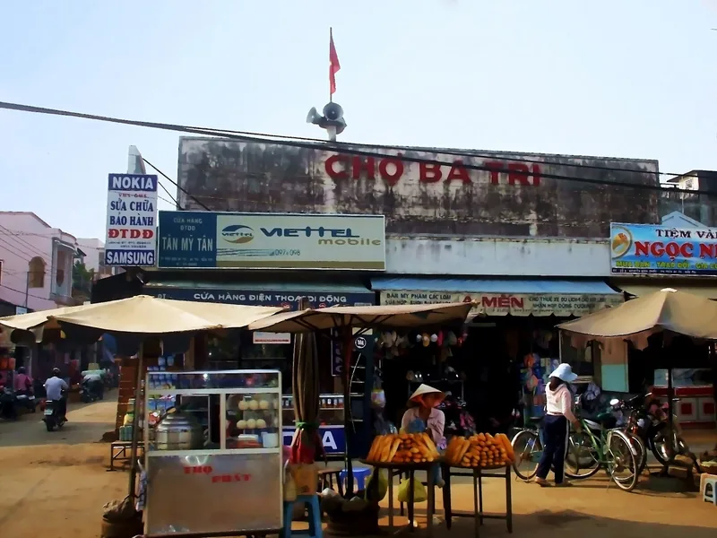 Người bán trái cây bên ngoài chợ Ba Trị với các bảng hiệu cửa hàng và xe đạp ở Đồng bằng sông Cửu Long.