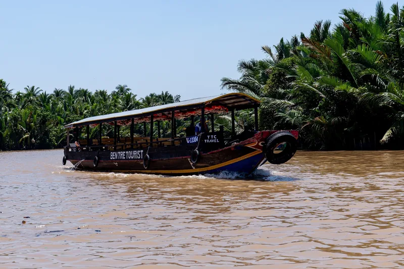 Một chiếc thuyền du lịch bằng gỗ chạy trên sông nước đục, hai bên là rừng dừa xanh ở vùng Bến Tre, đồng bằng sông Mekong.
