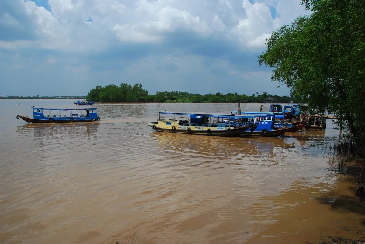 Những chiếc thuyền du lịch neo trên sông Mekong đục màu gần Tân Thạch, Bến Tre, với bờ sông xanh mát dưới trời nhiều mây.
