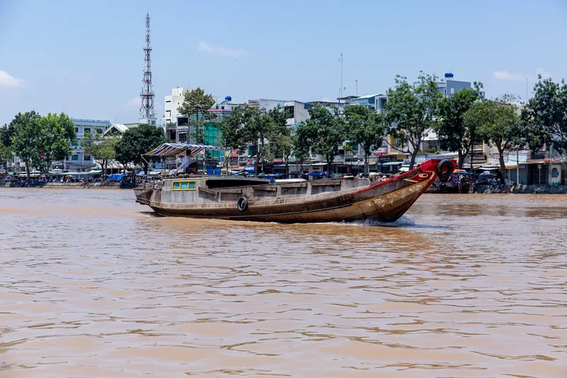 Một chiếc thuyền gỗ chở hàng truyền thống với mắt thuyền sơn màu lướt trên sông Mekong Delta gần khu nhà bên bờ sông.