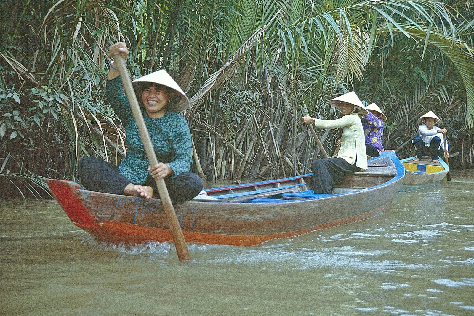 Những phụ nữ đội nón lá tươi cười chèo thuyền gỗ qua con rạch rợp bóng cây dừa nước ở Đồng bằng sông Cửu Long.