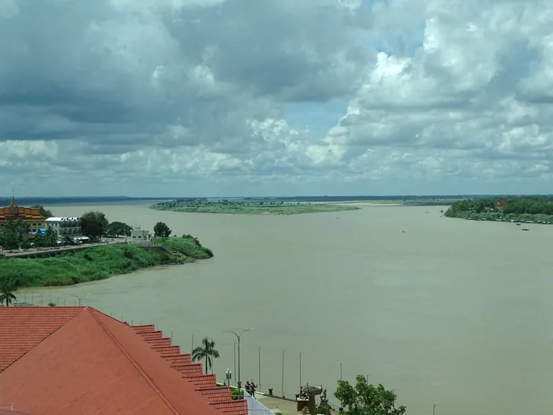 Toàn cảnh sông Mekong rộng lớn, nước đục màu phù sa uốn quanh khu nhà ven sông dưới bầu trời nhiều mây.
