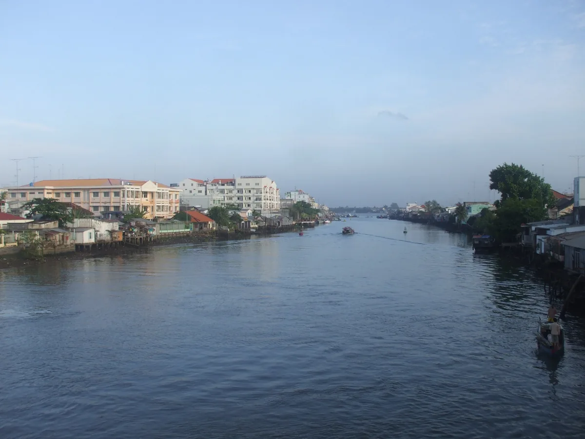 Dòng sông rộng chảy qua những căn nhà và tòa nhà ven sông dưới bầu trời buổi sáng nhạt màu ở thành phố Vĩnh Long.